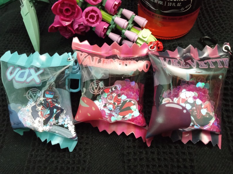 Puede incluir: Tres peque&ntilde;as bolsas de pl&aacute;stico transparente con bordes rosas, azules y morados. Cada bolsa tiene un nombre diferente impreso en la parte delantera: "Vox", "Valentino" y "Velvette". Cada bolsa contiene un peque&ntilde;o amuleto de personaje y purpurina de colores.