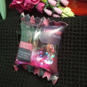Puede incluir: Una bolsa rosa y negra con una ventana de pl&aacute;stico transparente llena de purpurina rosa e iridiscente. La bolsa tiene el texto "LOVE OTI BY VELVET" y "TRY" impreso en ella. La bolsa tambi&eacute;n tiene el texto "PRODUCT DETAILS" y "THE BACKBONE OF THE VEES IS #THATBITCH" impreso en ella.