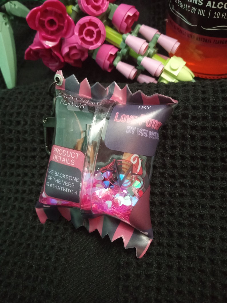 Puede incluir: Una bolsa rosa y negra con una ventana de pl&aacute;stico transparente llena de purpurina rosa e iridiscente. La bolsa tiene el texto "LOVE OTI BY VELVET" y "TRY" impreso en ella. La bolsa tambi&eacute;n tiene el texto "PRODUCT DETAILS" y "THE BACKBONE OF THE VEES IS #THATBITCH" impreso en ella.