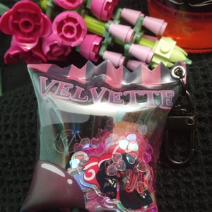 Puede incluir: Una bolsa de pl&aacute;stico transparente con un borde rayado rosa y negro y la palabra "VELVETTE" impresa en la parte delantera. La bolsa contiene una variedad de purpurina rosa y plateada y una pegatina negra y roja con un personaje de dibujos animados.