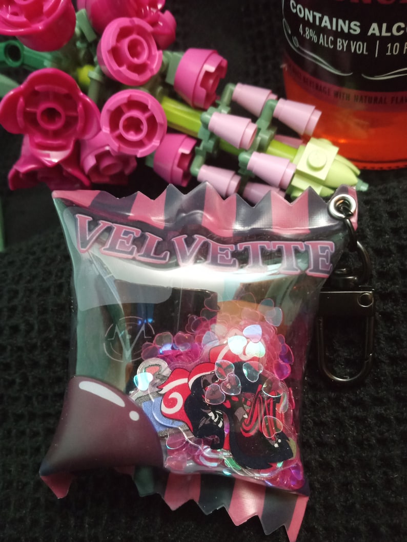 Puede incluir: Una bolsa de pl&aacute;stico transparente con un borde rayado rosa y negro y la palabra "VELVETTE" impresa en la parte delantera. La bolsa contiene una variedad de purpurina rosa y plateada y una pegatina negra y roja con un personaje de dibujos animados.