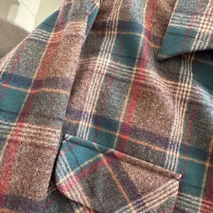 K&ouml;nnte beinhalten: Eine lang&auml;rmelige Jacke mit einem Karomuster in dunklem Petrol, Braun, Grau und d&uuml;nnen roten Linien. Das Kleidungsst&uuml;ck verf&uuml;gt &uuml;ber einen spitzen Kragen und eine sichtbare Pattentasche auf der Vorderseite aus einem strukturierten Stoff.