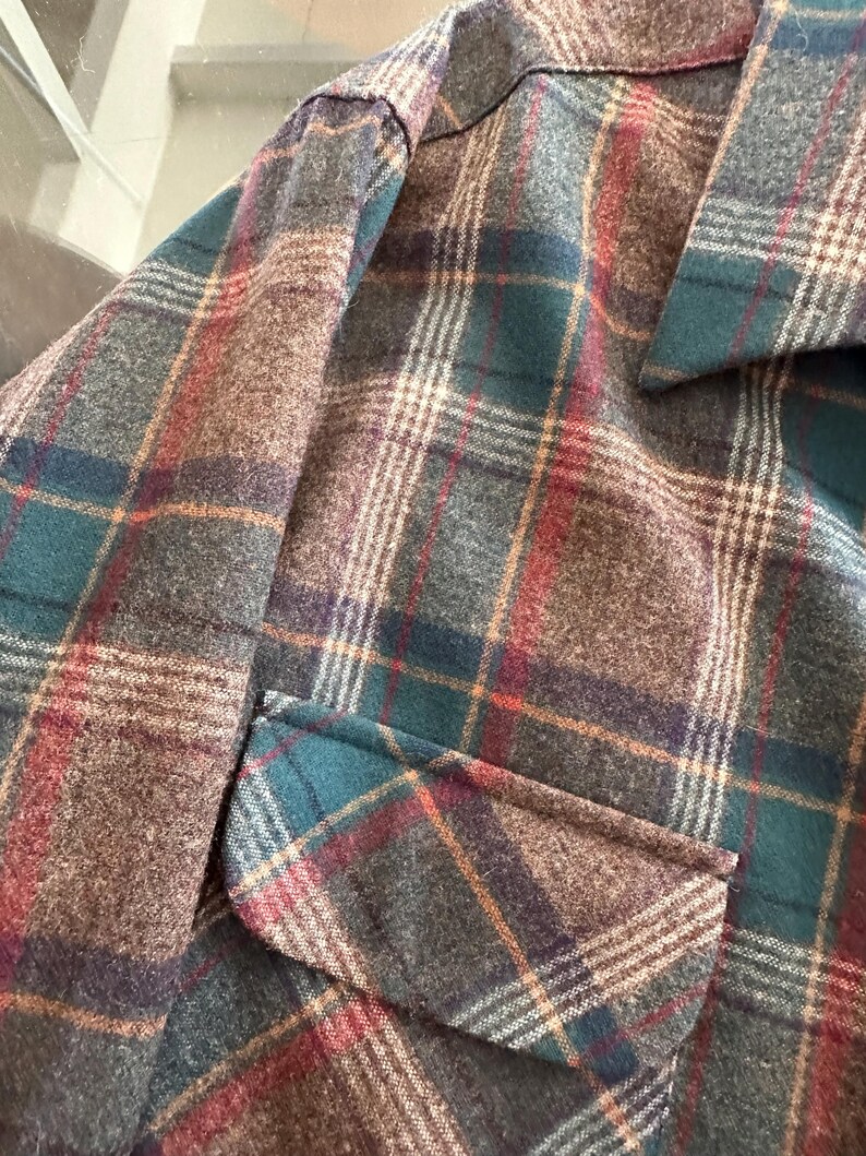 K&ouml;nnte beinhalten: Eine lang&auml;rmelige Jacke mit einem Karomuster in dunklem Petrol, Braun, Grau und d&uuml;nnen roten Linien. Das Kleidungsst&uuml;ck verf&uuml;gt &uuml;ber einen spitzen Kragen und eine sichtbare Pattentasche auf der Vorderseite aus einem strukturierten Stoff.