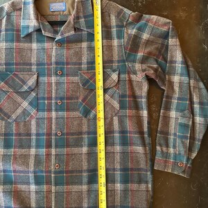 K&ouml;nnte beinhalten: Ein lang&auml;rmeliges Hemd mit Knopfleiste und einem Karomuster in Braun, Petrol und Rot. Das Hemd hat zwei Brusttaschen mit Klappen und ein Pendleton-Markenetikett am Kragen. Ein gelbes Ma&szlig;band zeigt eine L&auml;nge von etwa 76 cm an.