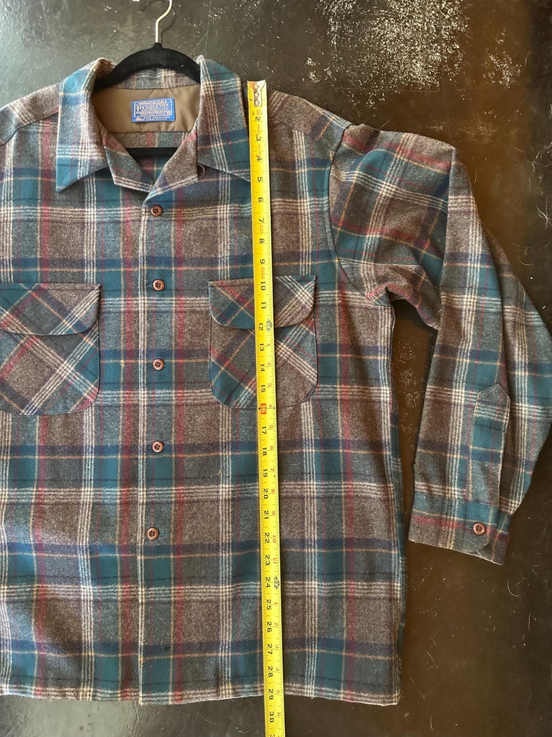 K&ouml;nnte beinhalten: Ein lang&auml;rmeliges Hemd mit Knopfleiste und einem Karomuster in Braun, Petrol und Rot. Das Hemd hat zwei Brusttaschen mit Klappen und ein Pendleton-Markenetikett am Kragen. Ein gelbes Ma&szlig;band zeigt eine L&auml;nge von etwa 76 cm an.