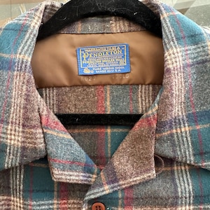 K&ouml;nnte beinhalten: Ein lang&auml;rmeliges Hemd mit Knopfleiste und einem Karomuster in Braun-, Petrol- und Rott&ouml;nen. Das Etikett am Innenkragen lautet: Warranted to be a Pendleton, Pendleton Woolen Mills, Portland, Oregon, 100% Virgin Wool, XL, Made in USA.