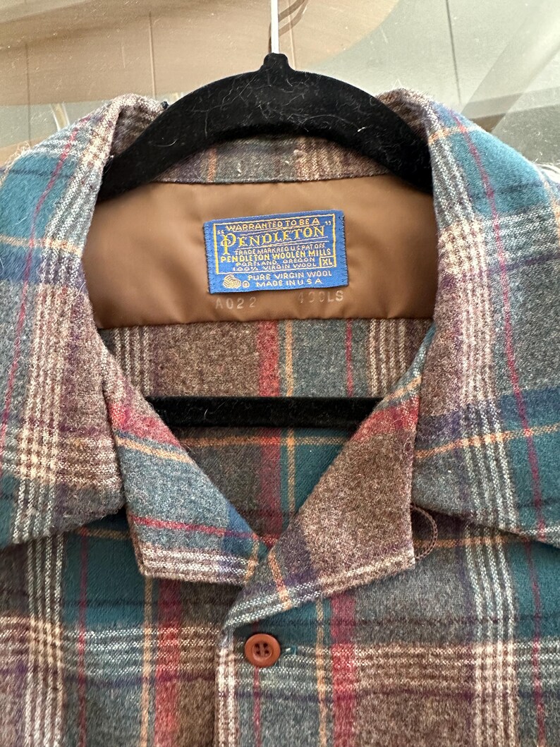 K&ouml;nnte beinhalten: Ein lang&auml;rmeliges Hemd mit Knopfleiste und einem Karomuster in Braun-, Petrol- und Rott&ouml;nen. Das Etikett am Innenkragen lautet: Warranted to be a Pendleton, Pendleton Woolen Mills, Portland, Oregon, 100% Virgin Wool, XL, Made in USA.