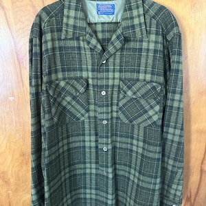 May include: A long-sleeve button-up shirt in a dark olive and forest green plaid pattern. The shirt features a pointed collar, two chest pockets with flaps, and a Pendleton brand label sewn into the inner neckline. The fabric has a textured finish.
