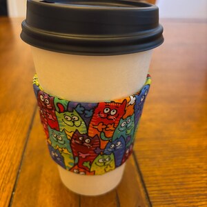 May include: A white paper coffee cup with a black lid and a colorful cat-themed sleeve. The sleeve features a repeating pattern of cartoon cats in various colors, including red, green, blue, and orange.