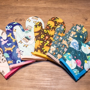 May include: Six colorful oven mitts with different animal and cartoon designs. The mitts are made of quilted fabric and have a pink, blue, or beige trim.