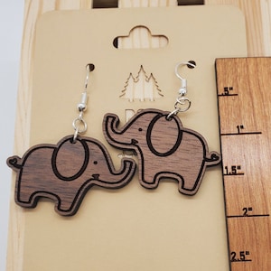 May include: A pair of wooden elephant earrings with silver hooks. The earrings are approximately 2 inches in length. The earrings are displayed on a wooden background with a ruler for size reference. The ruler is marked in inches and reads "PineStreetPorch Creations".
