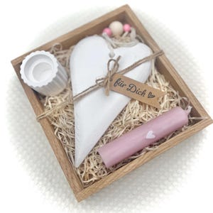 Könnte beinhalten: Eine Holzkiste mit dekorativen Gegenständen. Sie enthält ein weißes herzförmiges Ornament, eine rosa Kerze mit einem weißen Herzen, einen kleinen weißen Kerzenhalter und ein Etikett mit der Aufschrift "für Dich". Die Kiste ist mit Stroh gefüllt.