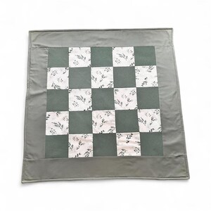 Könnte beinhalten: Ein quadratischer, handgefertigter Quilt mit Schachbrettmuster. Abwechselnd Quadrate sind einfarbiges, gedämpftes Grün und weißer Stoff mit zartem Blumendruck. Der Quilt ist von einem breiten, einfarbigen grauen Stoff umrandet.