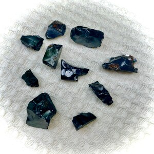 Könnte beinhalten: Eine Sammlung von zehn schwarzen Obsidian-Steinen in verschiedenen Formen und Größen. Die Steine sind auf einer weißen, strukturierten Oberfläche angeordnet.