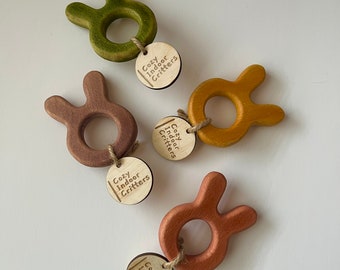 May include: Four wooden bunny-shaped teethers in different colors: green, brown, yellow, and orange. Each teether has a small wooden tag attached with the text "Cozy Indoor Critters".