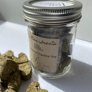 May include: A glass jar filled with green heart-shaped Timothy hay treats for small pets. The jar has a brown label that reads "Critter Treats Cozy Indoor Critters Strawberry Timothy Hay".