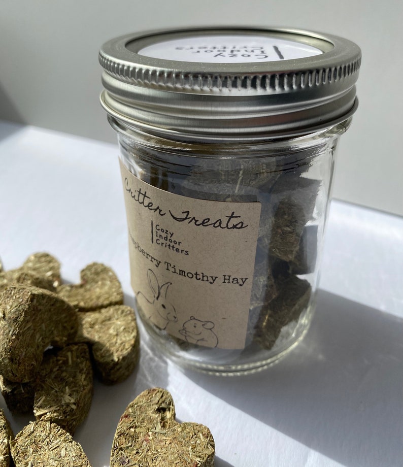 May include: A glass jar filled with green heart-shaped Timothy hay treats for small pets. The jar has a brown label that reads "Critter Treats Cozy Indoor Critters Strawberry Timothy Hay".
