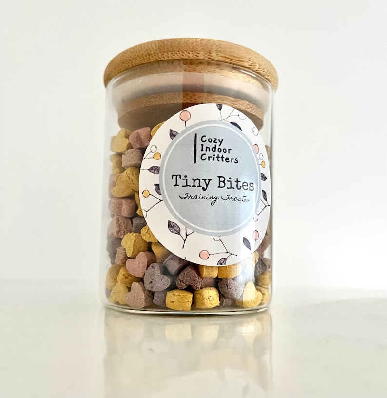 May include: A glass jar with a bamboo lid filled with heart-shaped training treats for small pets. The label reads "Cozy Indoor Critters Tiny Bites Training Treats".