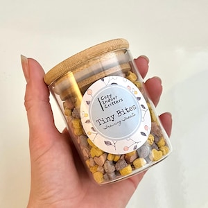 May include: A glass jar with a bamboo lid filled with small, colorful training treats. The jar has a white label with the text "Cozy Indoor Critters Tiny Bites Training Treats".
