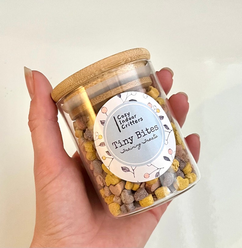 May include: A glass jar with a bamboo lid filled with small, colorful training treats. The jar has a white label with the text "Cozy Indoor Critters Tiny Bites Training Treats".