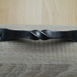 Könnte beinhalten: Ein schwarzer, handgeschmiedeter Eisen-Schrankgriff mit einem gedrehten Mittelteil. Der Griff ist rechteckig mit abgerundeten Enden, die jeweils mit einer schwarzen Schraube befestigt sind. Diese rustikale Hardware verleiht Möbeln eine einzigartige Note.