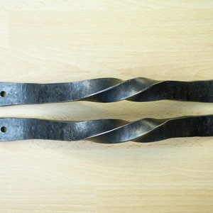 Könnte beinhalten: Zwei schwarze, gedrehte Türgriffe aus Metall mit gehämmerter Oberfläche. Die Griffe sind etwa 25 cm lang.