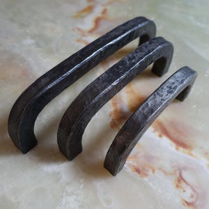 Könnte beinhalten: Drei schwarze Metall-Schrankgriffe mit gehämmerter Oberfläche. Die Griffe sind in verschiedenen Größen und in einer Reihe vom größten zum kleinsten angeordnet.