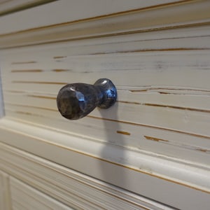 Könnte beinhalten: Nahaufnahme einer weißen Schranklade mit einer abgenutzten Oberfläche. Die Schublade hat einen gehämmerten Metallknopf in dunkelgrauer Farbe.