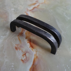 Könnte beinhalten: Zwei dunkelgraue, gebogene Metallgriffe. Die Griffe haben ein rustikales, geschmiedetes Aussehen und sind wahrscheinlich für einen Schrank oder eine Schublade. Sie liegen auf einer hellen, marmorierten Oberfläche.
