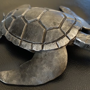 Könnte beinhalten: Eine handgefertigte Metallschildkröten-Skulptur mit einer strukturierten, dunkelgrauen Oberfläche. Die Schildkröte hat einen detaillierten Panzer, Flossen und einen Kopf und zeigt eine rustikale, handwerkliche Ästhetik. Geeignet für die Inneneinrichtung.
