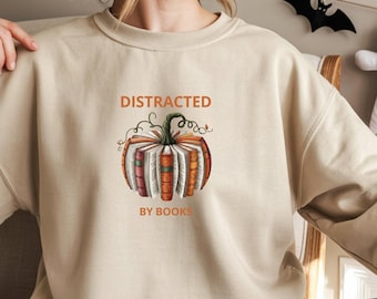 May include: Sand-colored sweatshirt with the text "DISTRACTED BY BOOKS" above a pumpkin illustration made of books. The sweatshirt is a neutral color and has long sleeves. The design is in shades of orange, brown, and white.