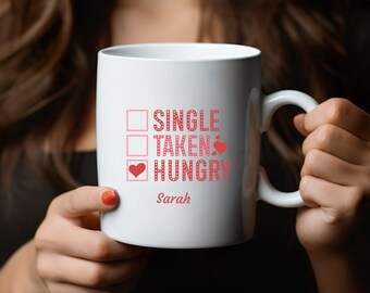 Può includere: Tazza in ceramica bianca con un cuore rosso e il testo "Single, Taken, Hungry" con un segno di spunta accanto a "Hungry". Il nome "Sarah" è stampato sotto il testo.