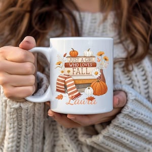 Op de afbeelding: Witte keramische mok met een herfstthema. De mok heeft pompoenen, boeken, een sjaal en de tekst "JUST A GIRL WHO LOVES FALL" en een naam. Het ontwerp bevat oranje, witte en bruine kleuren.