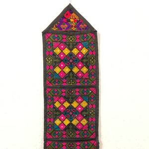 May include: A black fabric wall organizer with three pockets. Each pocket is decorated with colorful embroidery and mirror accents. The pockets are arranged vertically.