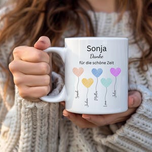 May include: White ceramic mug with the name "Sonja" and the German phrase "Danke f&uuml;r die sch&ouml;ne Zeit." The mug features four colourful heart-shaped balloons with names below. The mug is held by a person wearing a cream-coloured sweater.