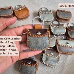 May include: Handmade leather keychains in various colors, including gray and brown. Each keychain features a metal ring, snap button closure, and orange stitching. The text "100% Handmade" is visible.