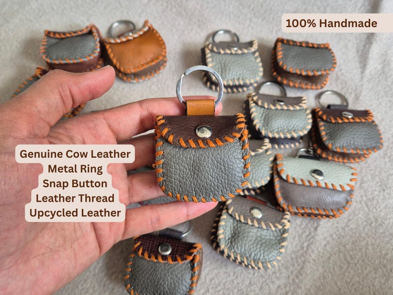May include: Handmade leather keychains in various colors, including gray and brown. Each keychain features a metal ring, snap button closure, and orange stitching. The text "100% Handmade" is visible.