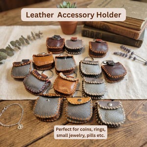 May include: A collection of small leather accessory holders in various colors, including brown, gray, and tan. Each holder has a stitched border and a metal keyring. The text "Leather Accessory Holder" is at the top. The text "Perfect for coins, rings, small jewelry, pills etc." is at the bottom.