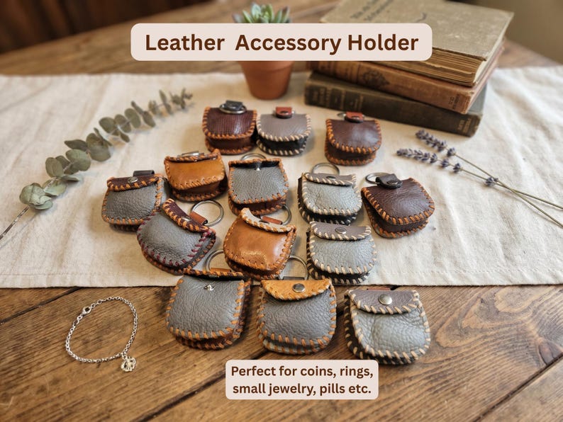 May include: A collection of small leather accessory holders in various colors, including brown, gray, and tan. Each holder has a stitched border and a metal keyring. The text "Leather Accessory Holder" is at the top. The text "Perfect for coins, rings, small jewelry, pills etc." is at the bottom.