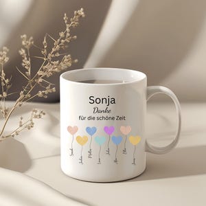 May include: White ceramic mug with the name "Sonja" and the phrase "Danke f&uuml;r die sch&ouml;ne Zeit" printed in black. The mug features colorful heart-shaped balloons with names below them. A sprig of dried flowers is in the background.