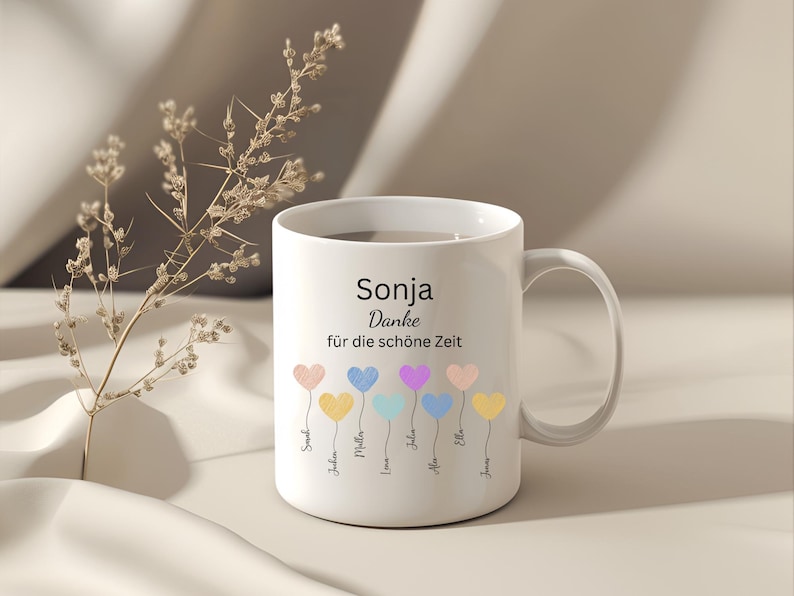 May include: White ceramic mug with the name "Sonja" and the phrase "Danke f&uuml;r die sch&ouml;ne Zeit" printed in black. The mug features colorful heart-shaped balloons with names below them. A sprig of dried flowers is in the background.