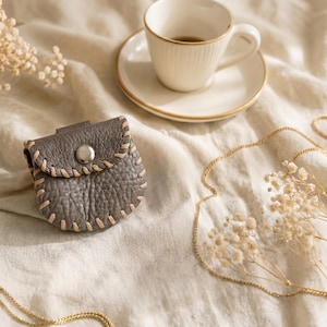 May include: A small, gray leather pouch with a button closure and beige stitching. The pouch is on a cream-colored surface, next to a teacup and saucer, a gold necklace, and dried flowers. The pouch is approximately 3 inches wide.