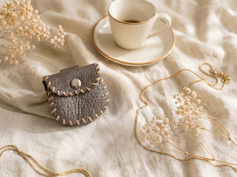 May include: A small, gray leather pouch with a button closure and beige stitching. The pouch is on a cream-colored surface, next to a teacup and saucer, a gold necklace, and dried flowers. The pouch is approximately 3 inches wide.