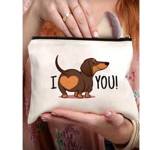 Peut inclure: Pochette zippée de couleur crème avec un teckel de dessin animé et un cœur sur l'arrière-train. Le texte "I YOU!" est imprimé sur la pochette. Un pinceau de maquillage est visible à l'intérieur.