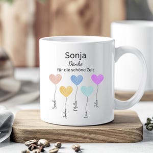 May include: White ceramic mug with a handle, featuring the name "Sonja" and the phrase "Danke f&uuml;r die sch&ouml;ne Zeit." Colorful heart-shaped balloons with names below them decorate the mug. The mug sits on a wooden surface.