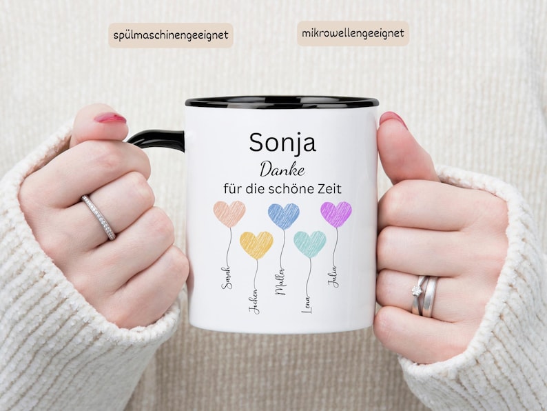May include: White ceramic mug with a black rim and handle, featuring the name "Sonja" and the phrase "Danke f&uuml;r die sch&ouml;ne Zeit." The mug has colourful heart-shaped balloons with names below. The mug is held by a person.