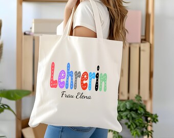May include: A white canvas tote bag with the text "Lehrerin Frau Elena" in colorful, dotted letters. The bag has two handles and is being held by a person.
