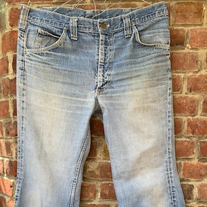 Peut inclure: Jean en denim bleu clair avec une jambe évasée. Le jean a une conception classique à cinq poches, une fermeture à bouton et à glissière et des passants de ceinture. Le denim a un aspect délavé, suggérant l'usure et l'âge.