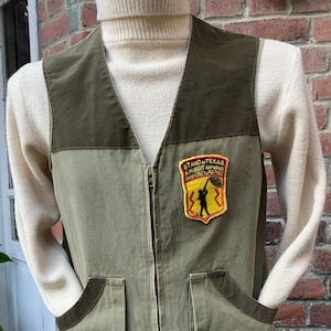 Peut inclure: Un gilet de chasse vert avec deux grandes poches et une fermeture éclair. Le gilet a un écusson jaune avec le texte "STAND & TEXAS A. HUBERT ARMURIER SUSI CREC'Y PARTNER" et une silhouette noire d'une personne tenant une arme à feu. Il y a aussi un écusson rouge, blanc et bleu avec le texte "CRAF".