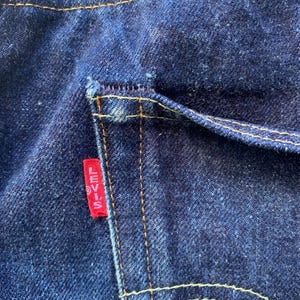 May include: Dark indigo blue denim jeans featuring a back pocket with thick, golden-yellow contrast stitching. A small, rectangular red fabric tag is sewn into the side of the pocket, clearly displaying the white text Levi's in capital letters.